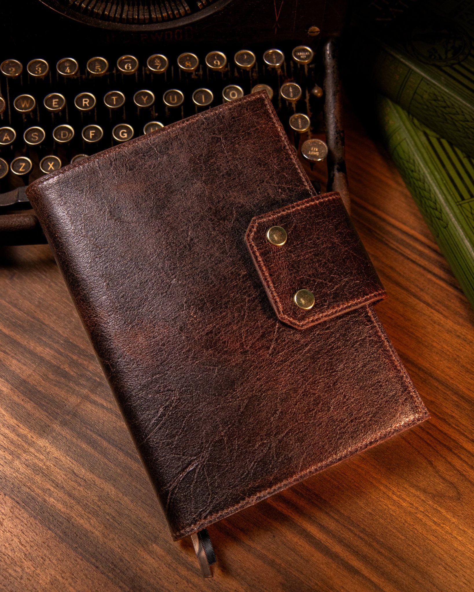 Brown leather notebook with gold closure on a wooden surface next to an old typewriter.