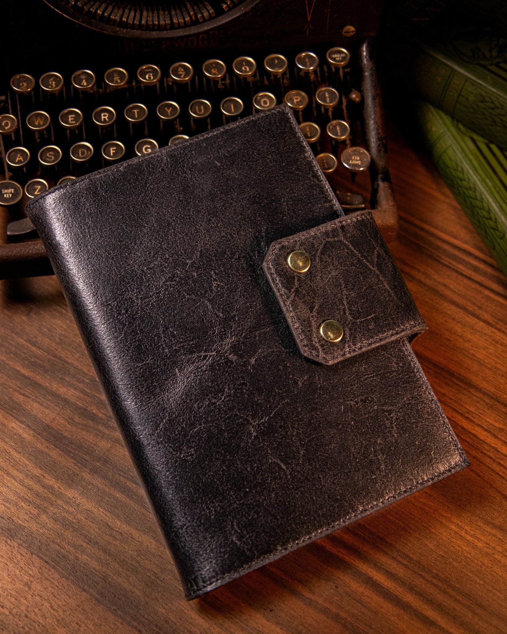 Black leather notebook with gold closure on a wooden surface with an old typewriter in the background.