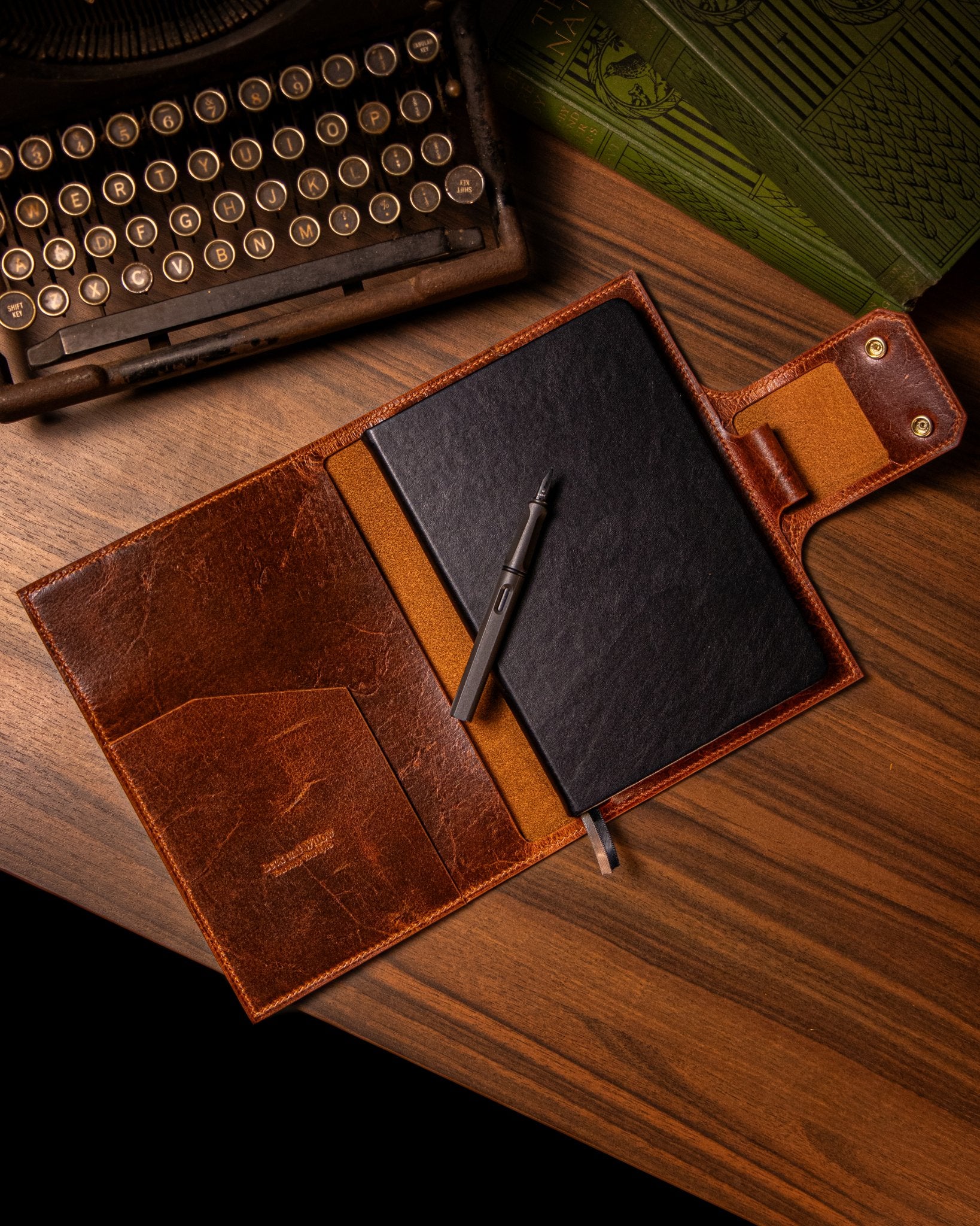 Tan leather journal with a pen and black notebook on a wooden surface, next to an old typewriter.
