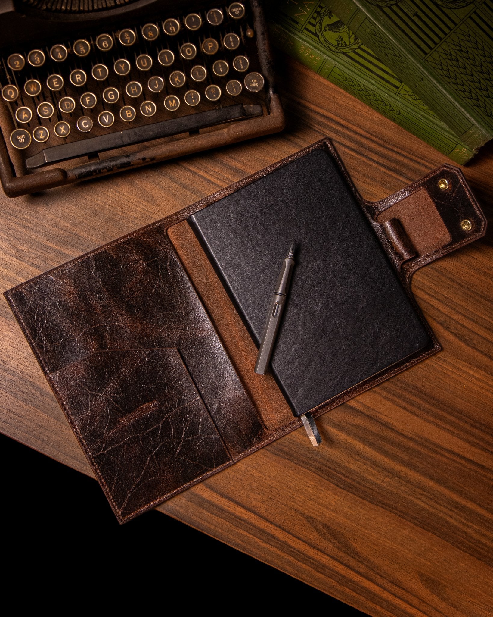 Brown leather journal with a pen and notebook on a wooden surface next to an old typewriter.