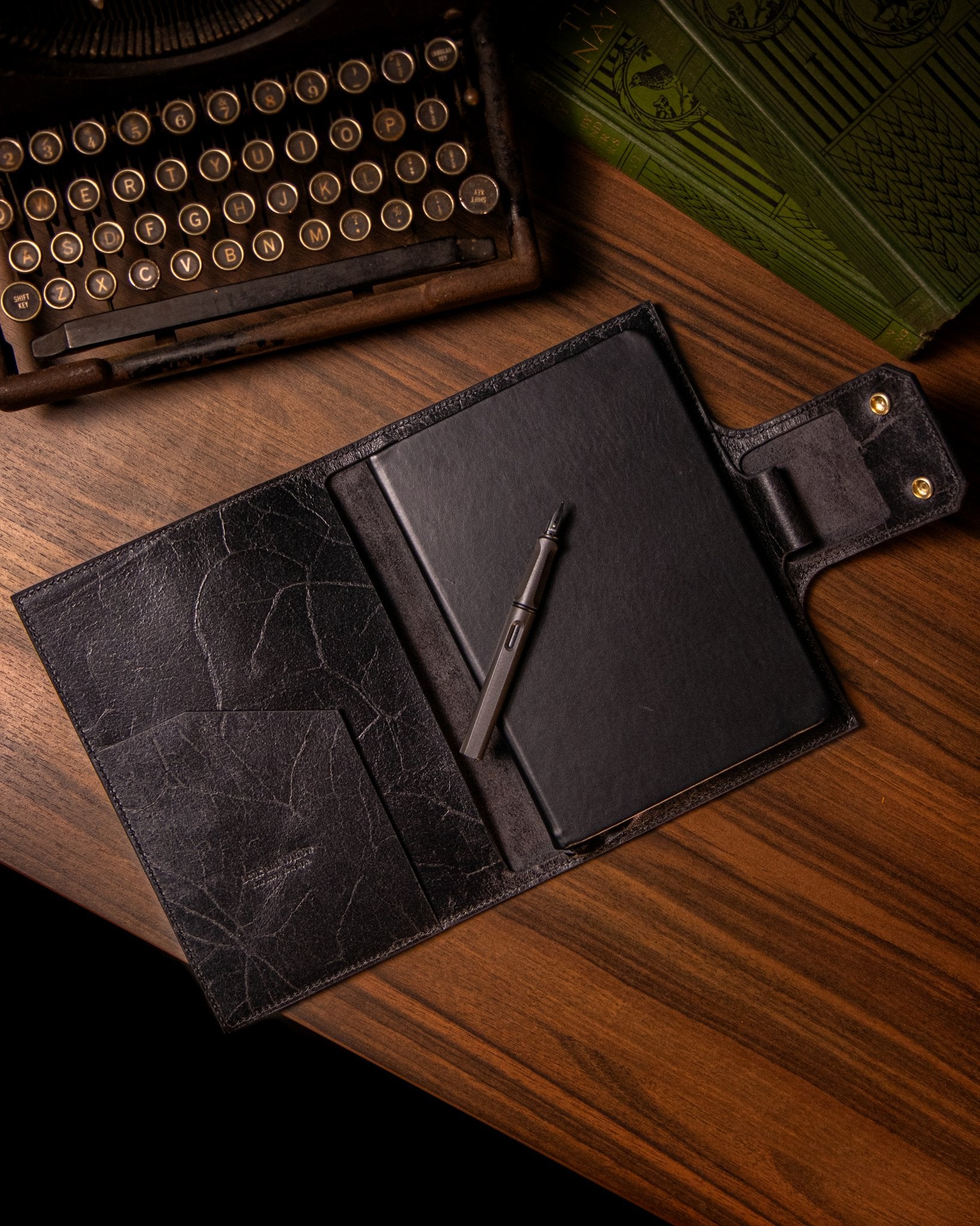 Black leather journal with a pen and notebook on a wooden surface next to an old typewriter.