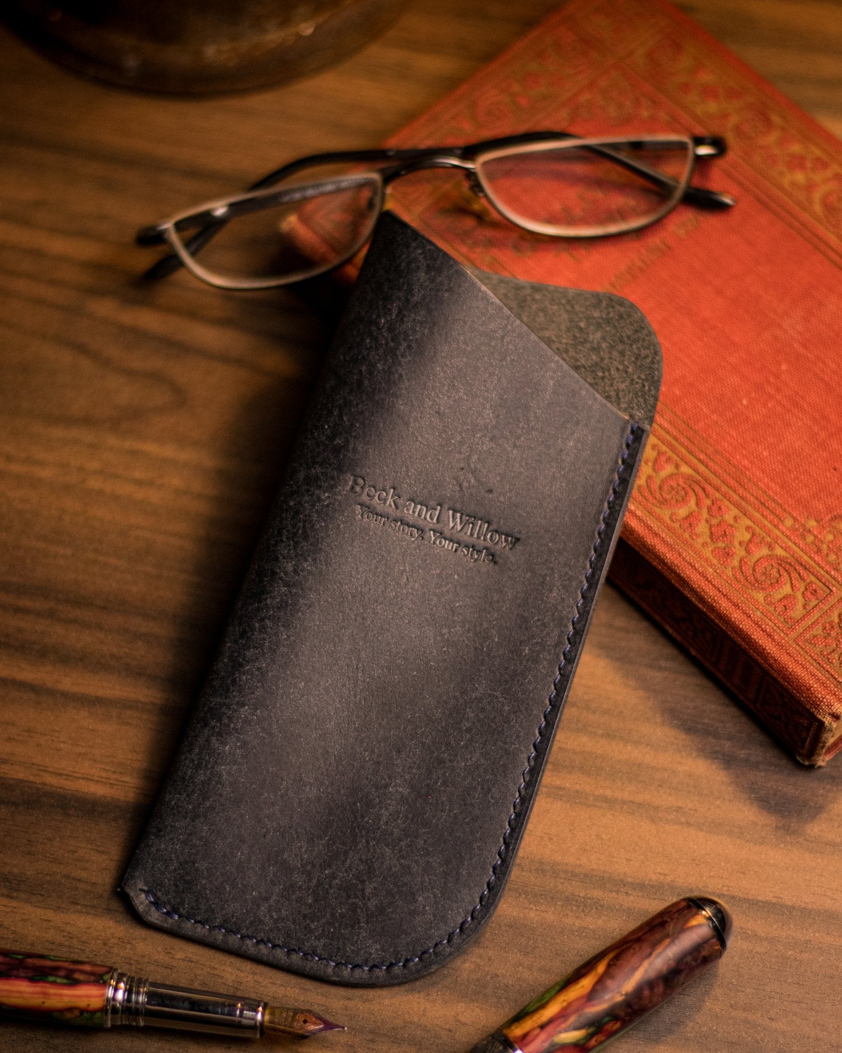 Lifestyle shot of an italian leather glasses case in navy with a book and glasses 