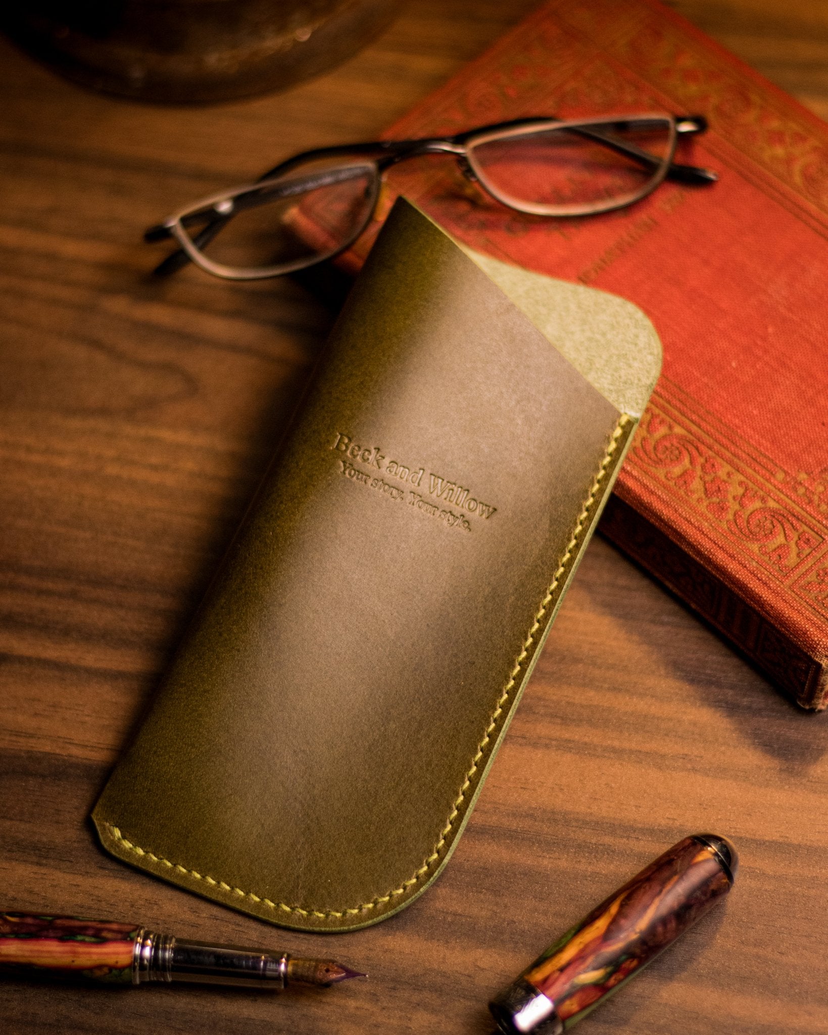 Lifestyle shot of an olive italian leather glasses case with a book and glasses