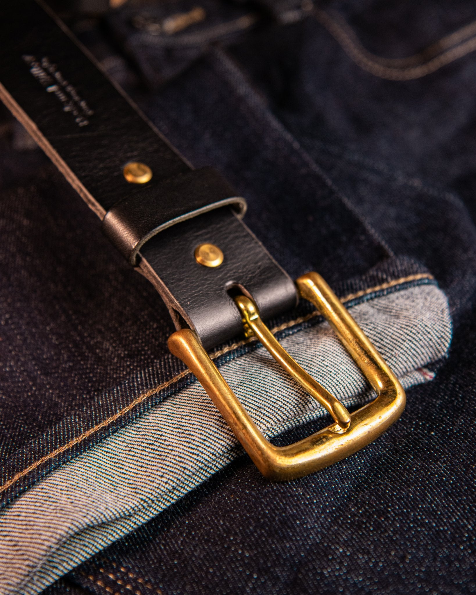 Close-up of a dark leather belt with a gold buckle on a denim background