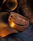 Brown leather belt with brass buckle on denim fabric, with brown boots in the background.