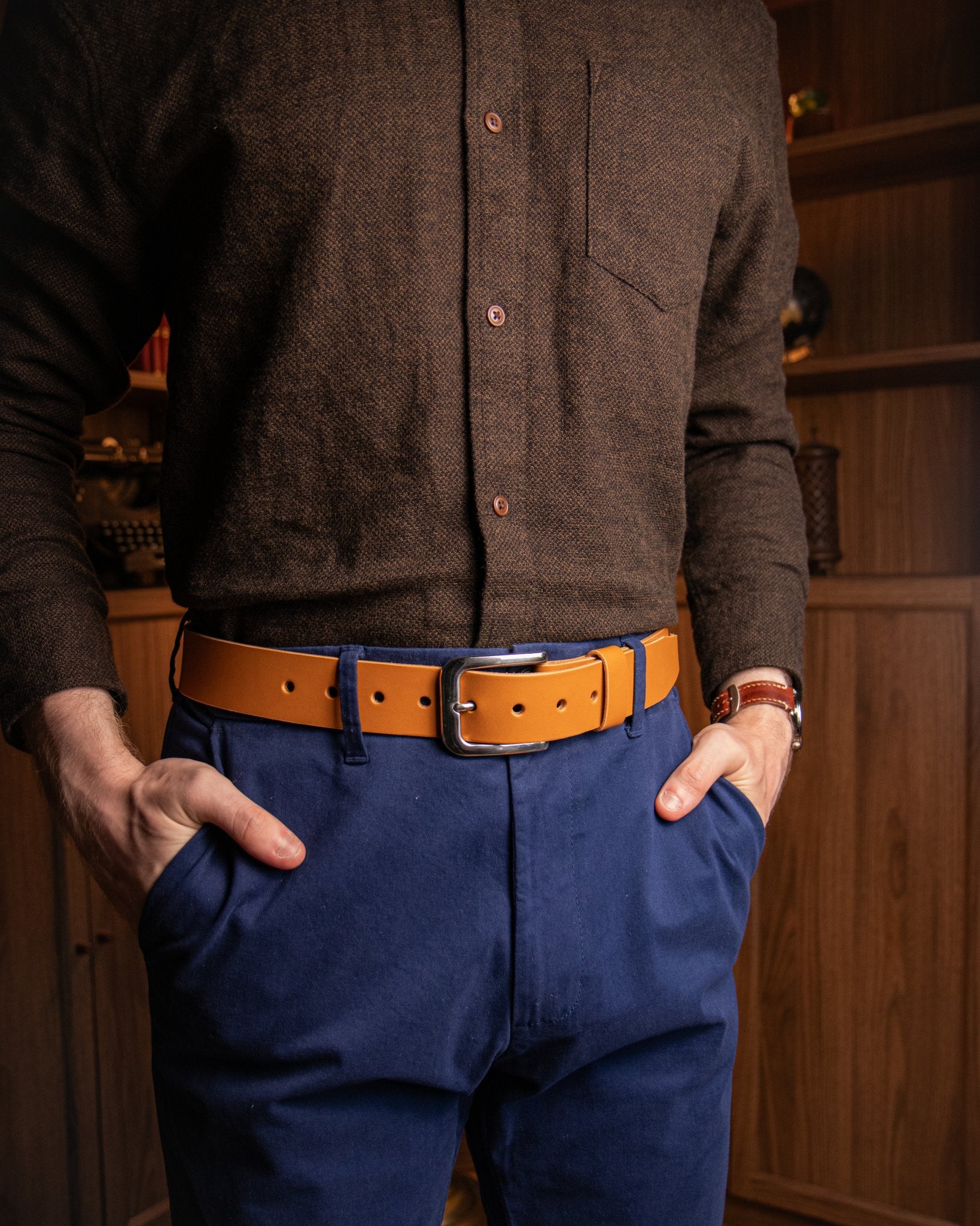 Person wearing a brown shirt and blue pants with a brown belt, standing against a wooden background.