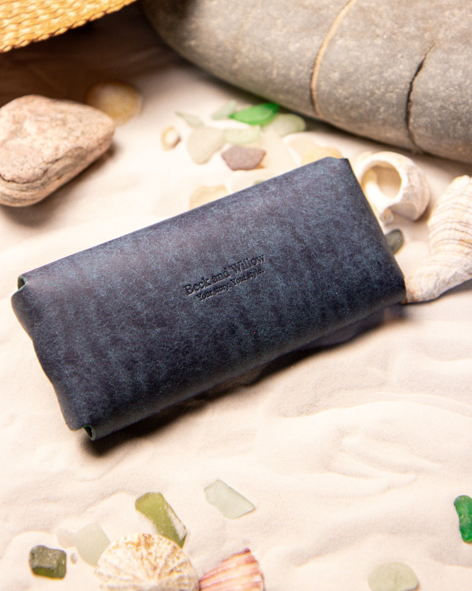 Blue eyewear pouch with 'Beck and Willow' branding on a sandy background with stones and shells.