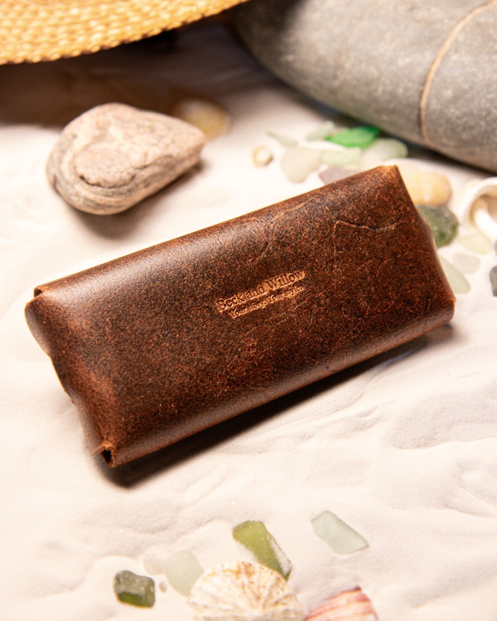Tan leather glasses pouch with 'Beck and Willow' branding on a textured surface with stones and shells.