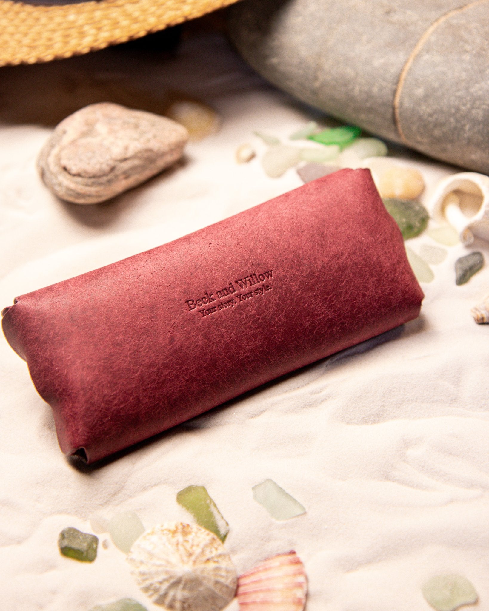 Red Leather sunglasses case with 'Beck and Willow' branding on a beach setting with stones and shells.