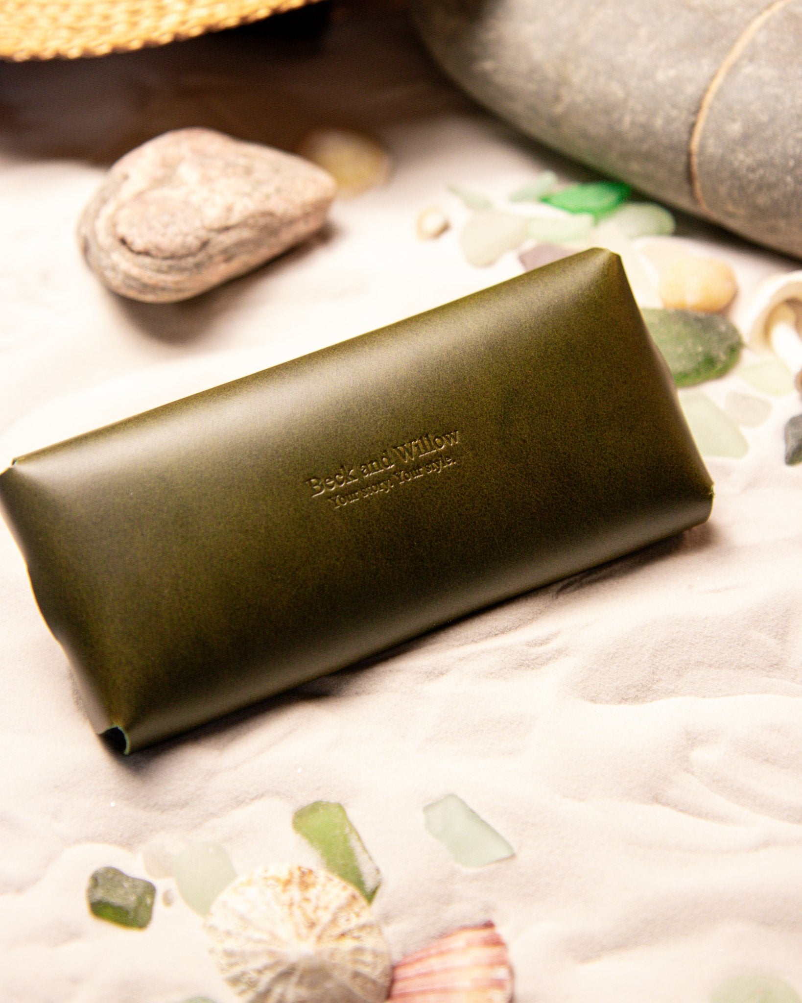 Olive green leather glasses case with 'Beck and Willow' branding on a beach setting with shells and stones.