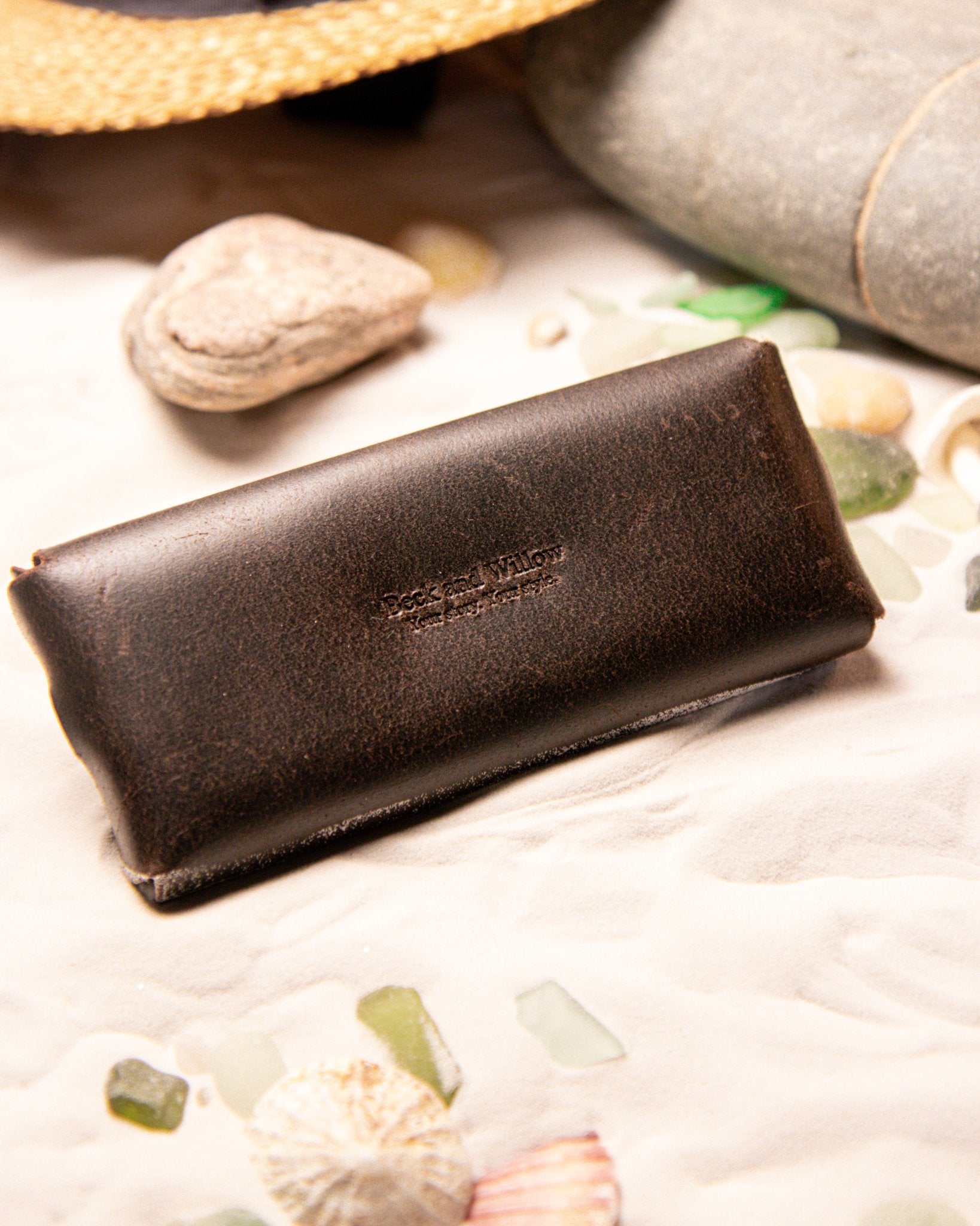 Brown leather glasses pouch on a beach with shells and rocks with branding 'Beck and Willow'