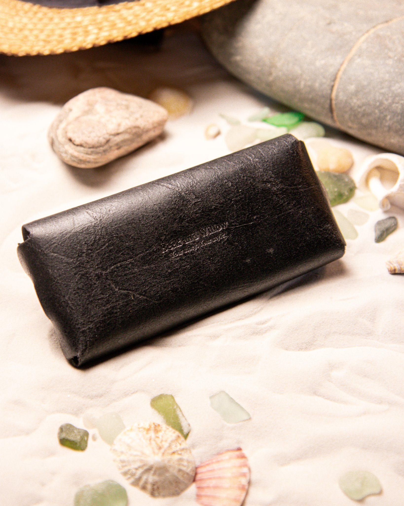 Black leather glasses case on a sandy surface with seashells and pebbles