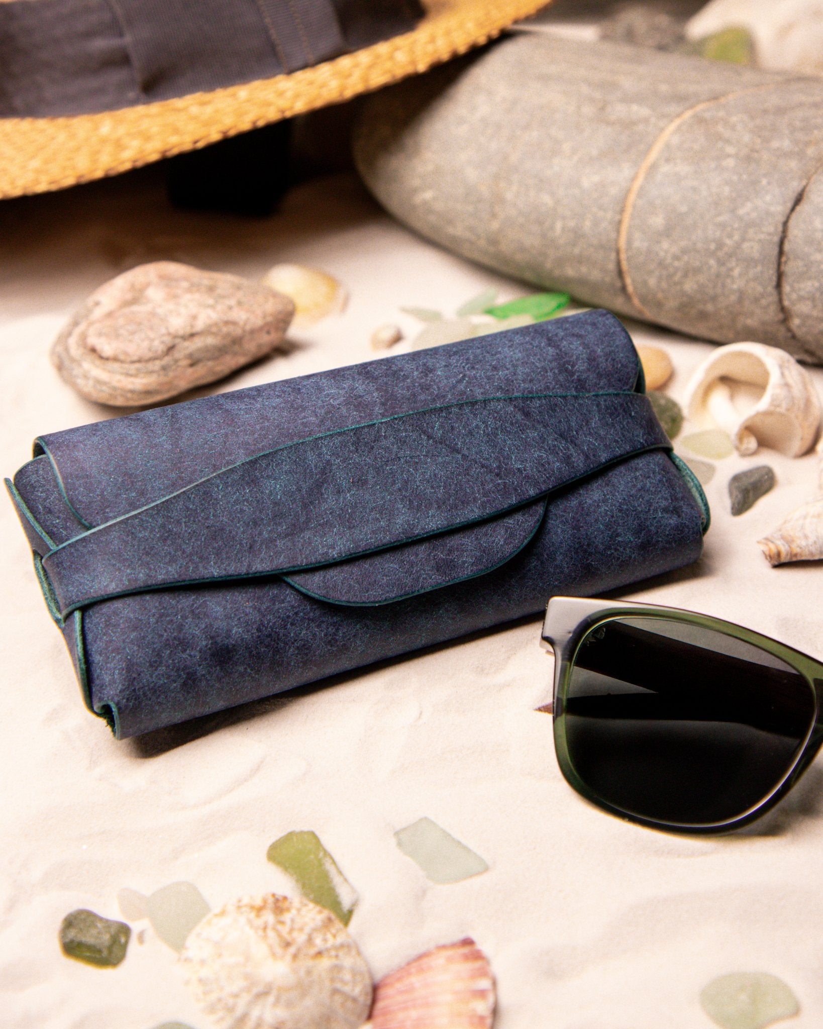 Blue leather sunglasses case and sunglasses on a sandy surface with seashells and rocks.