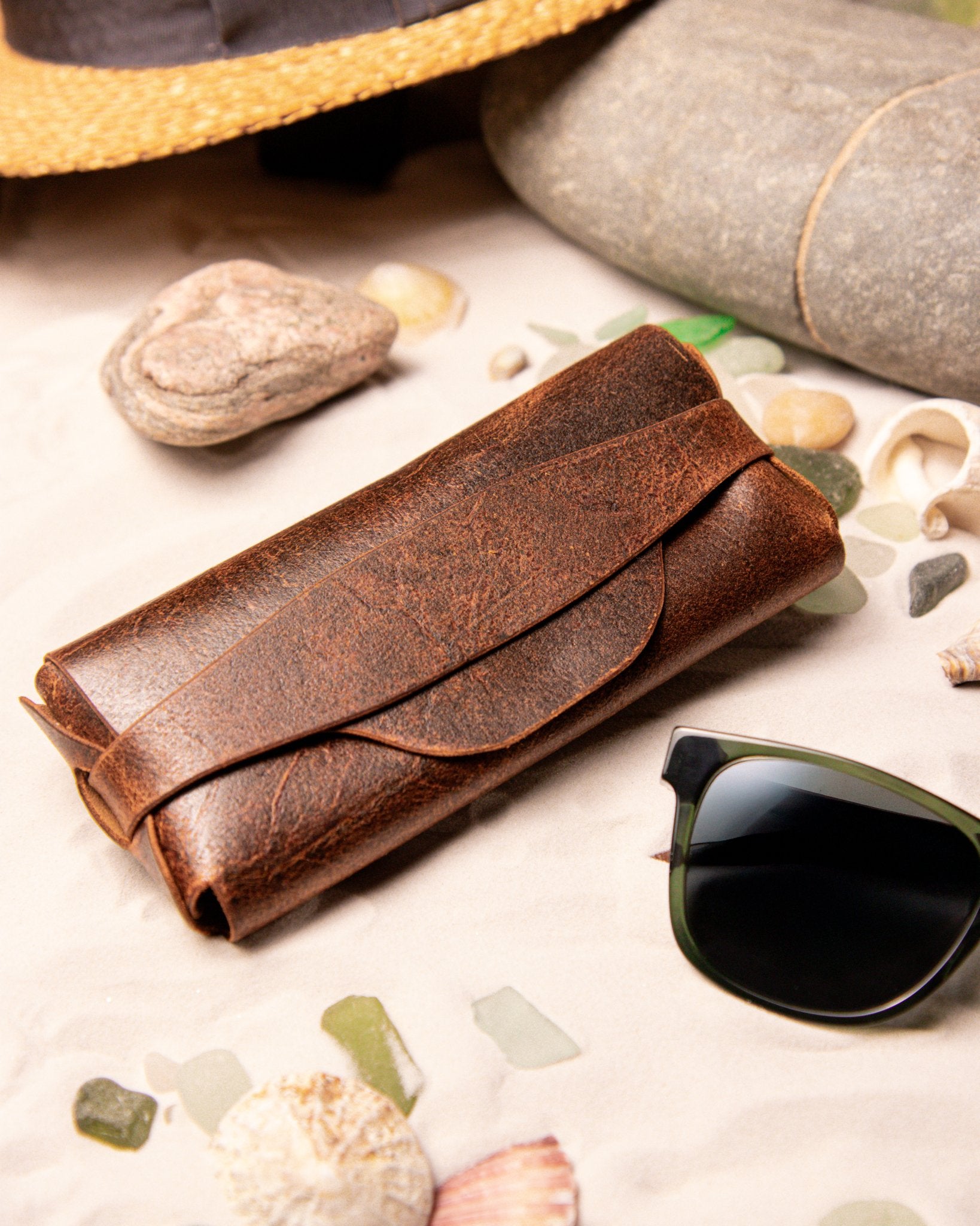 Tan leather sunglasses case on a surface with seashells and stones