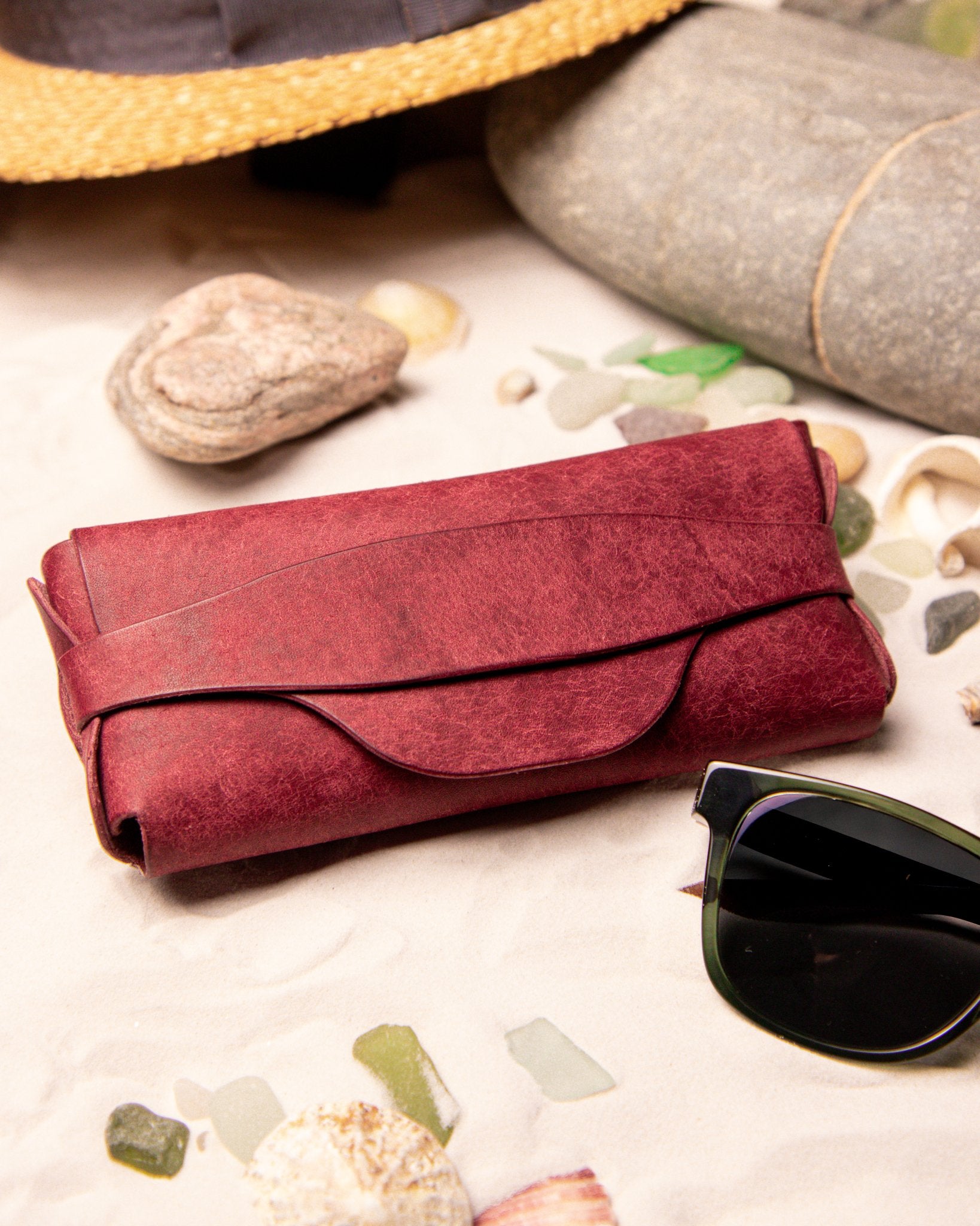 Red leather eyewear case on a sandy surface with sunglasses and seashells