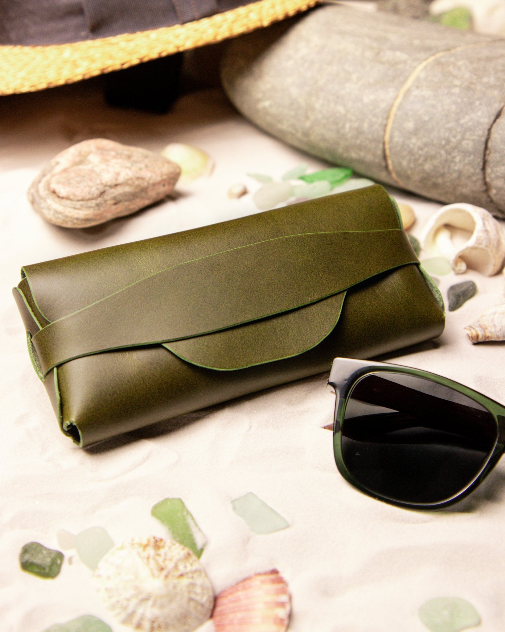 Olive green leather sunglasses case and sunglasses on a sandy surface with shells and pebbles.