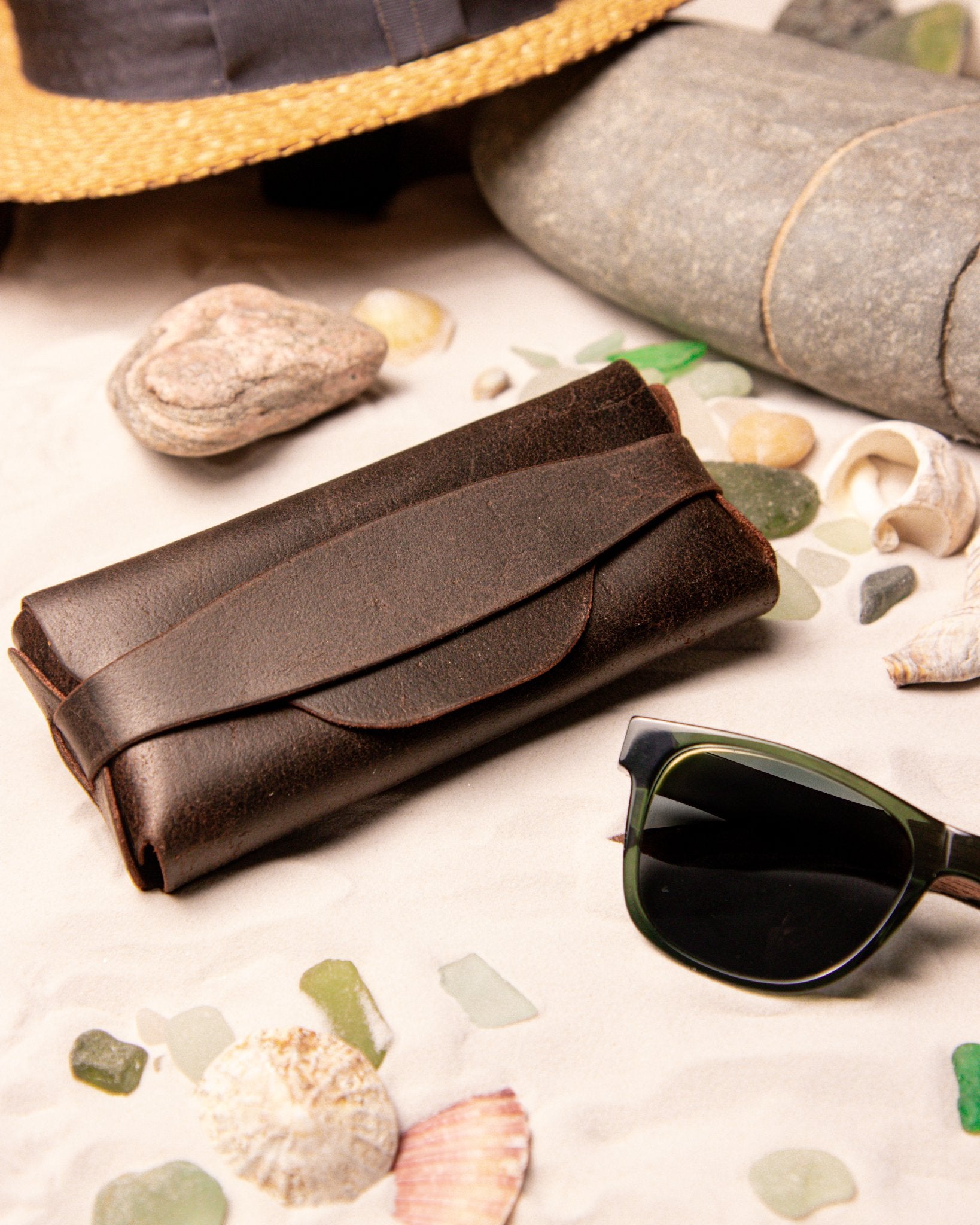 Brown leather eyewear case and sunglasses on a beach setting with shells and stones.
