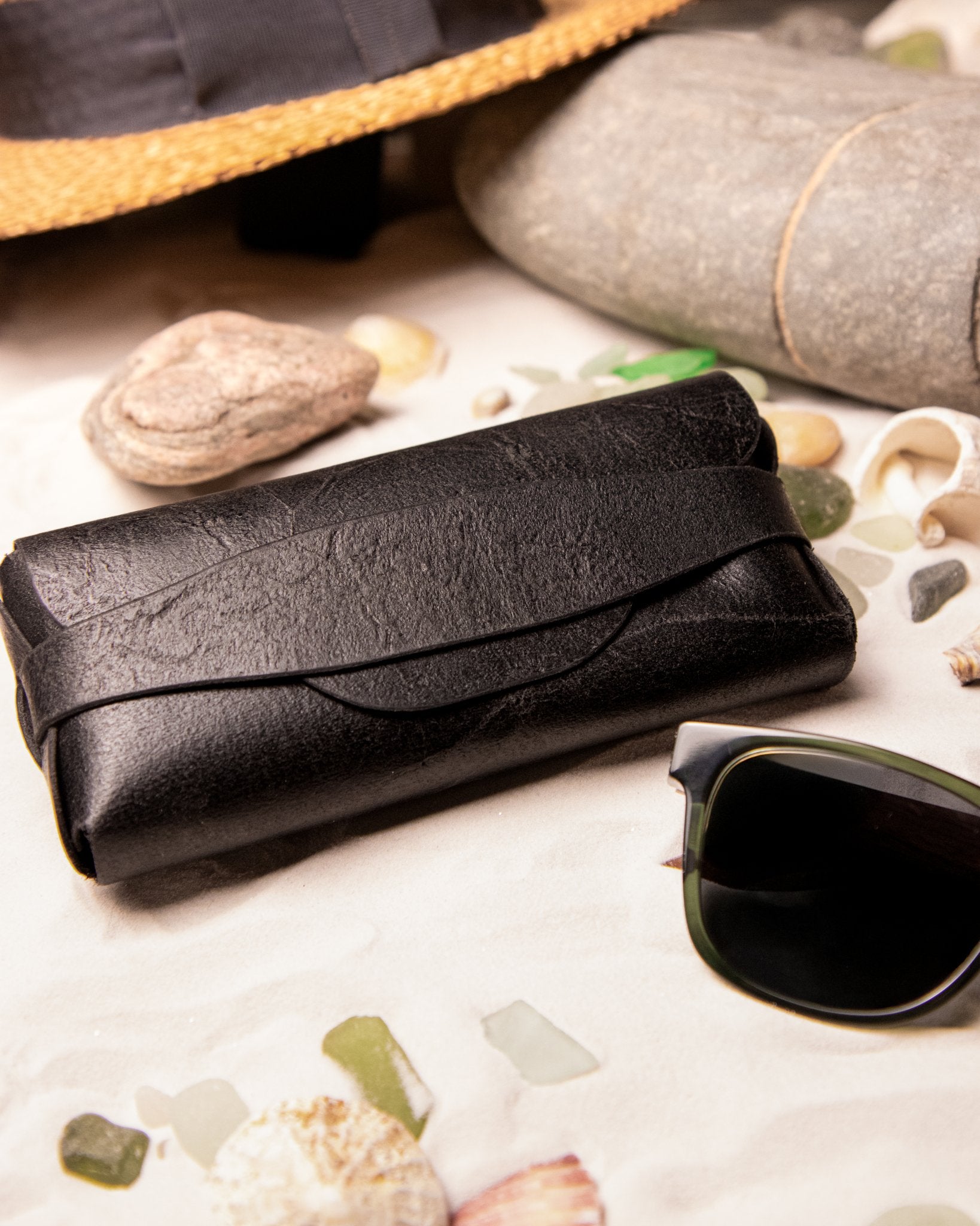 Black leather glasses pouch on a textured surface with sunglasses and seashells.