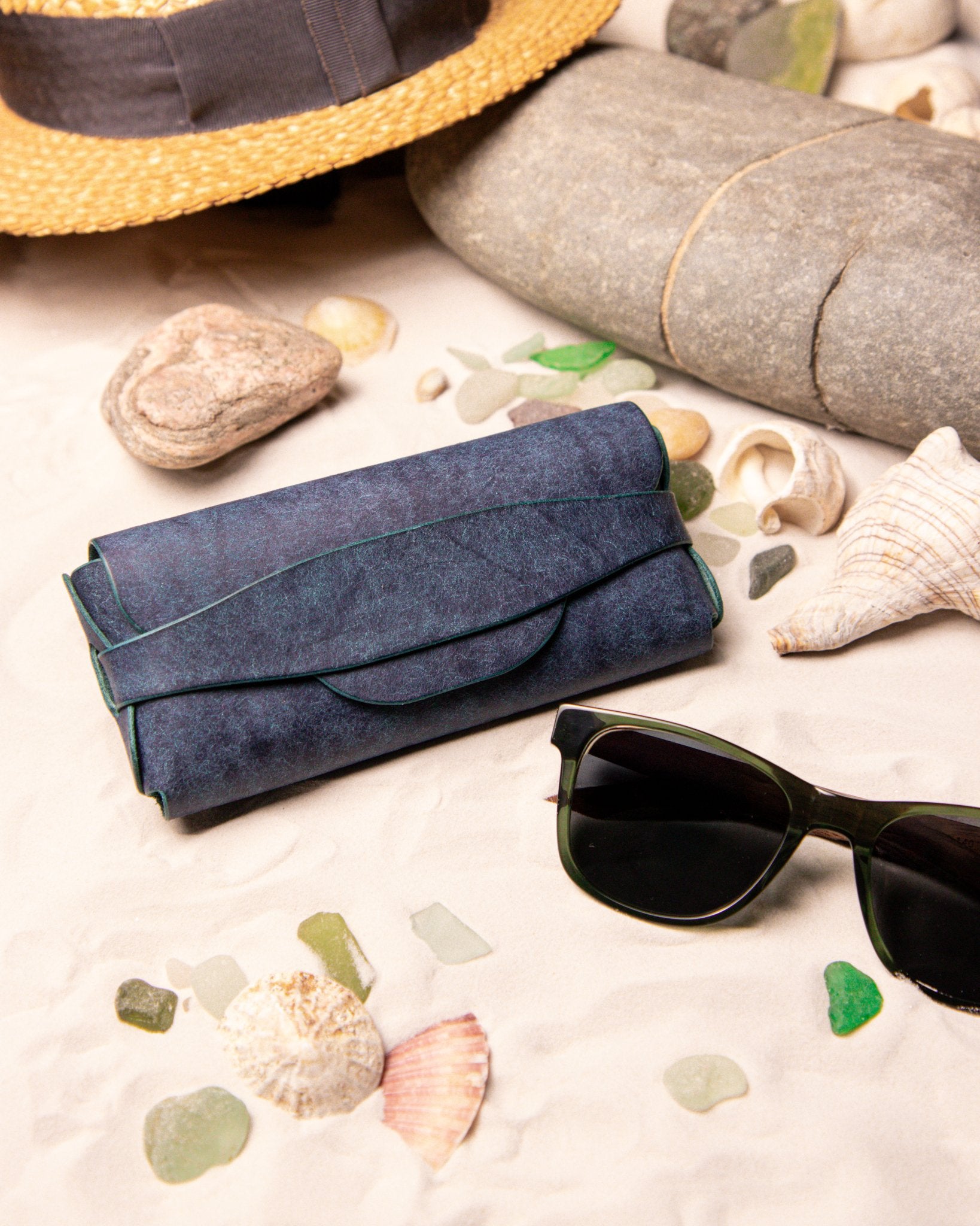 Blue leather glasses case with sunglasses on a sandy surface with seashells and stones.