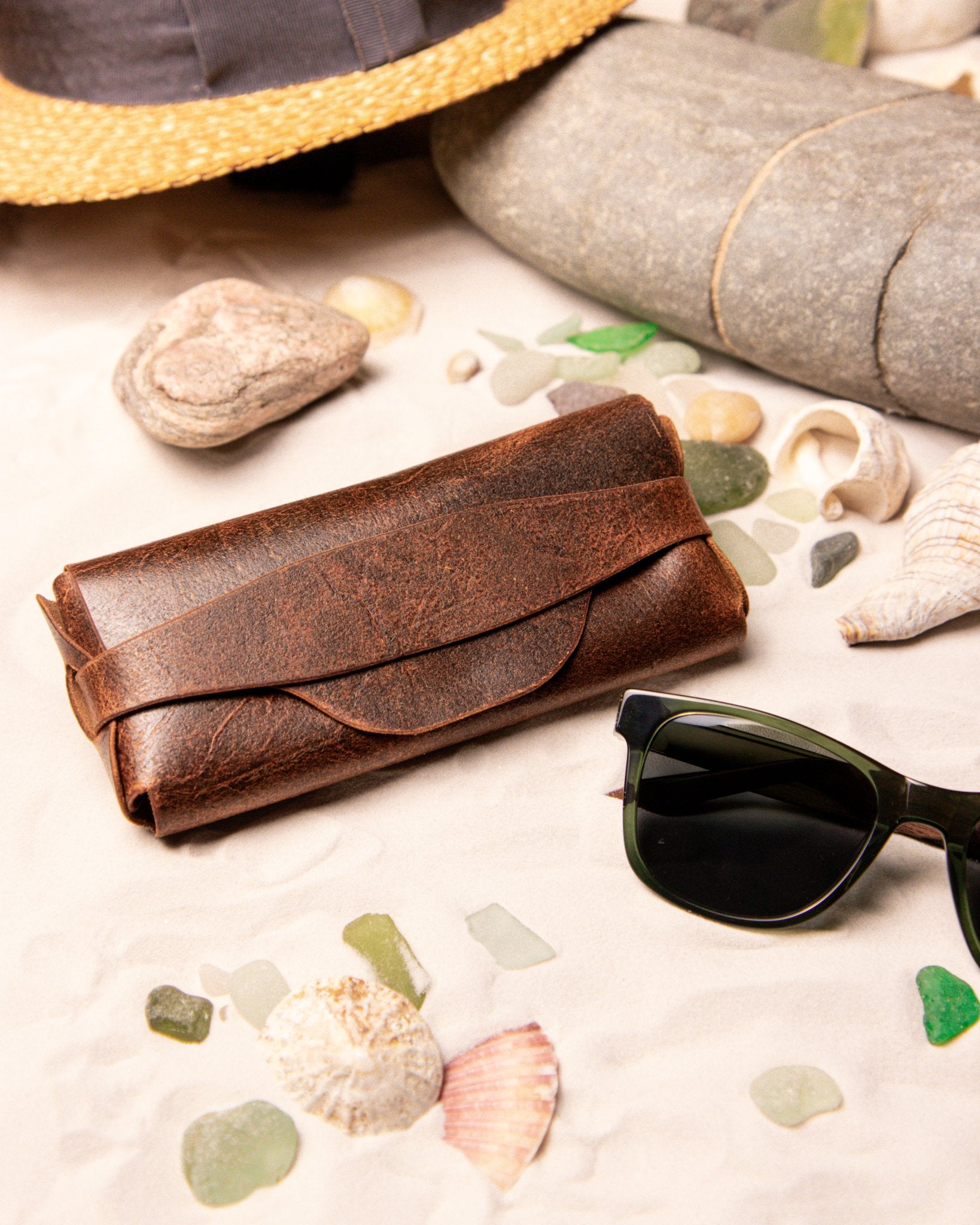Tan leather glasses case on a sandy surface with seashells and sunglasses.