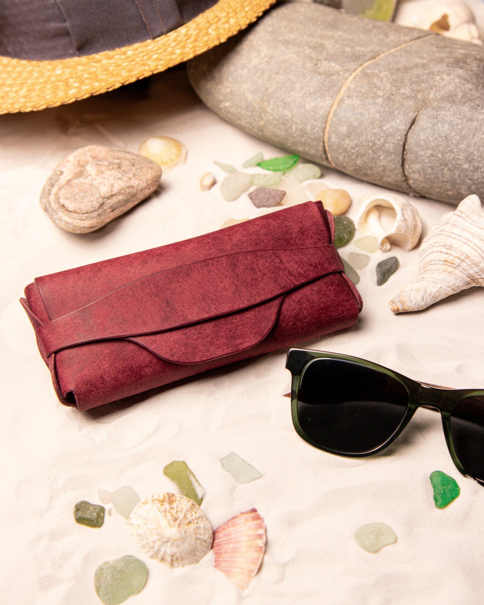 Burgundy Leather Sunglasses Case with sunglasses on a sandy surface with shells and pebbles.