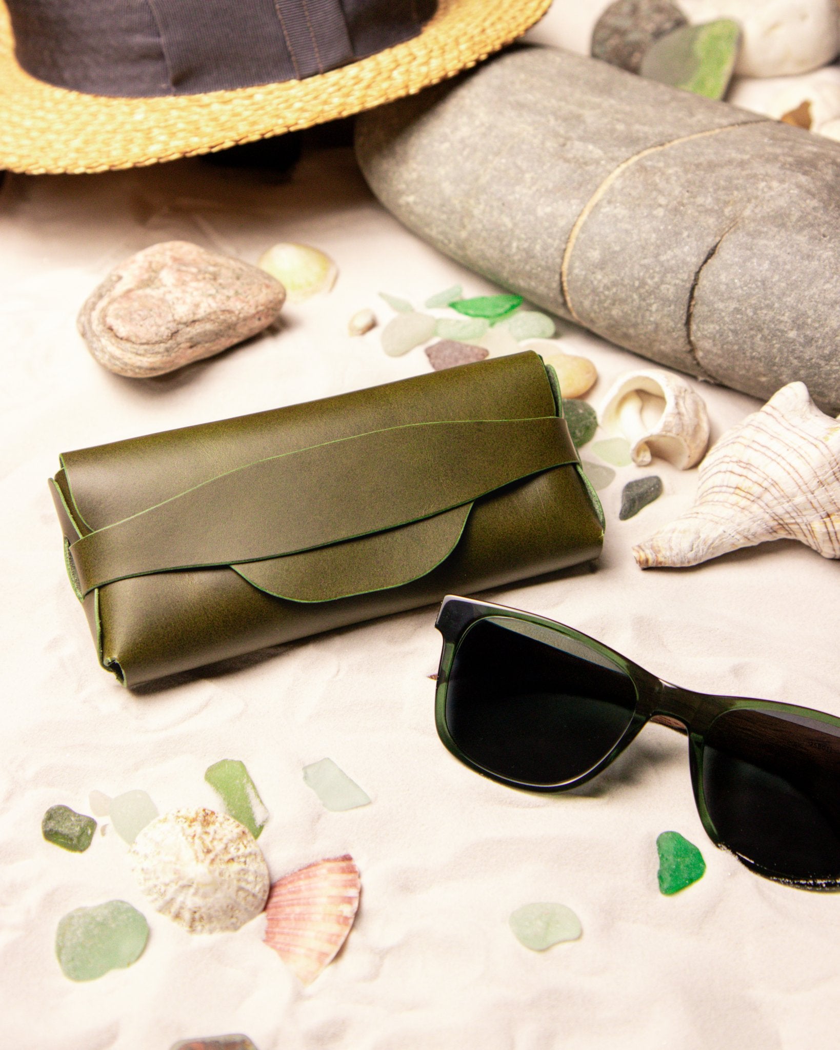 Olive green leather glasses case and sunglasses on a beach setting with shells and pebbles.
