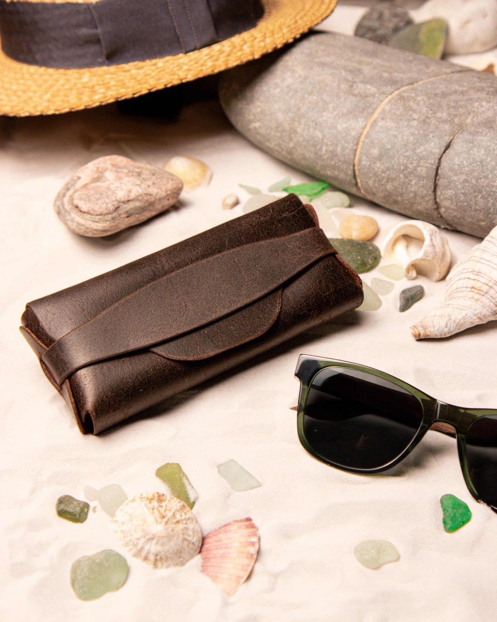 Brown leather glasses case, sunglasses, and straw hat on a sandy surface with shells and pebbles.