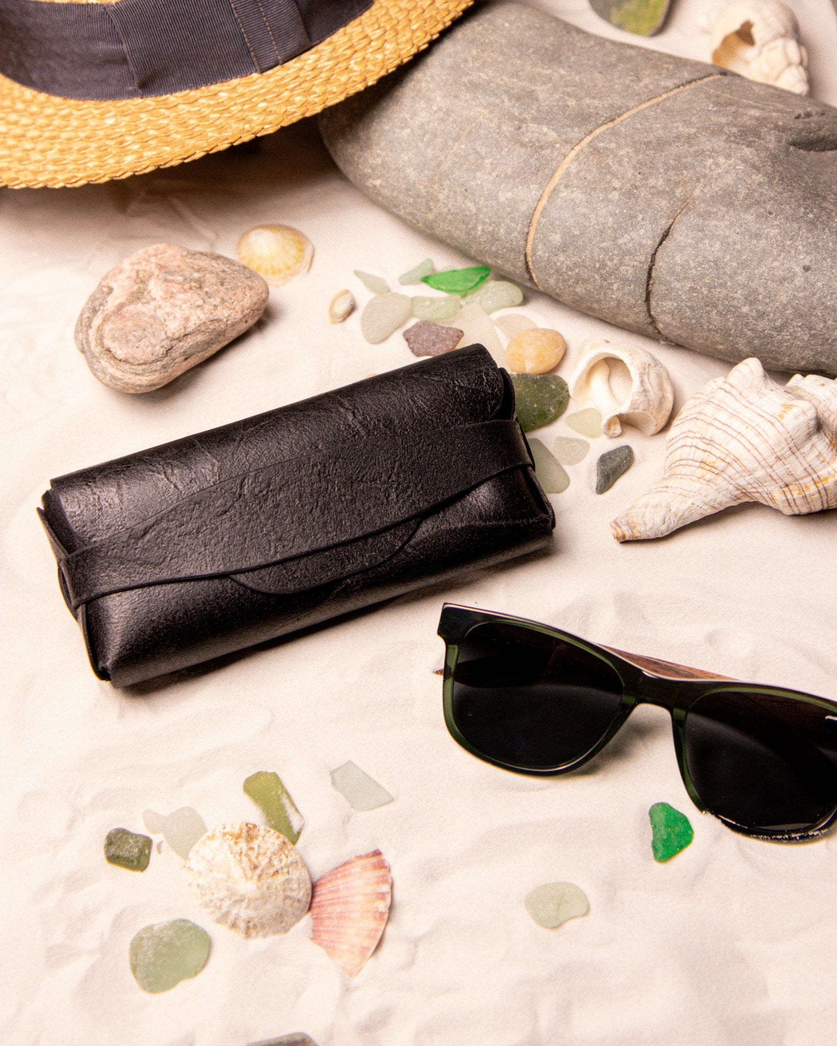 Black sunglasses and leather case on a sandy surface with seashells