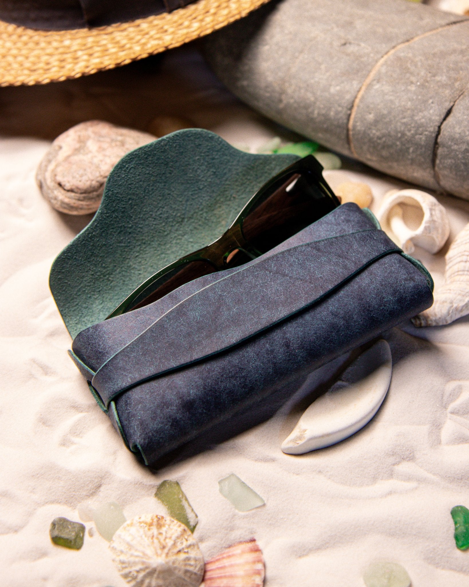 Blue leather eyewear case with sunglasses on a sandy surface with shells and stones.
