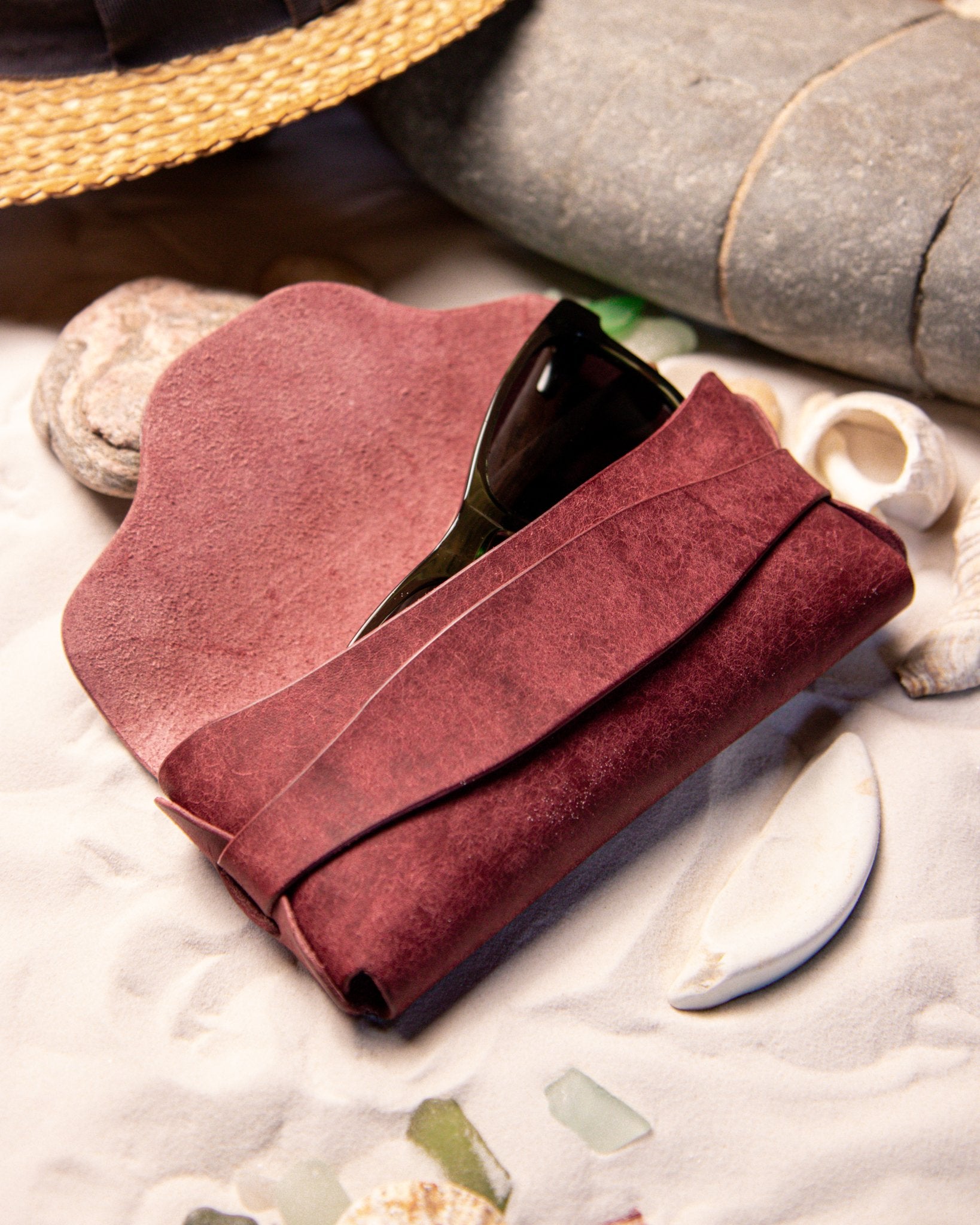 Red leather glasses case with sunglasses on a sandy surface with seashells and rocks