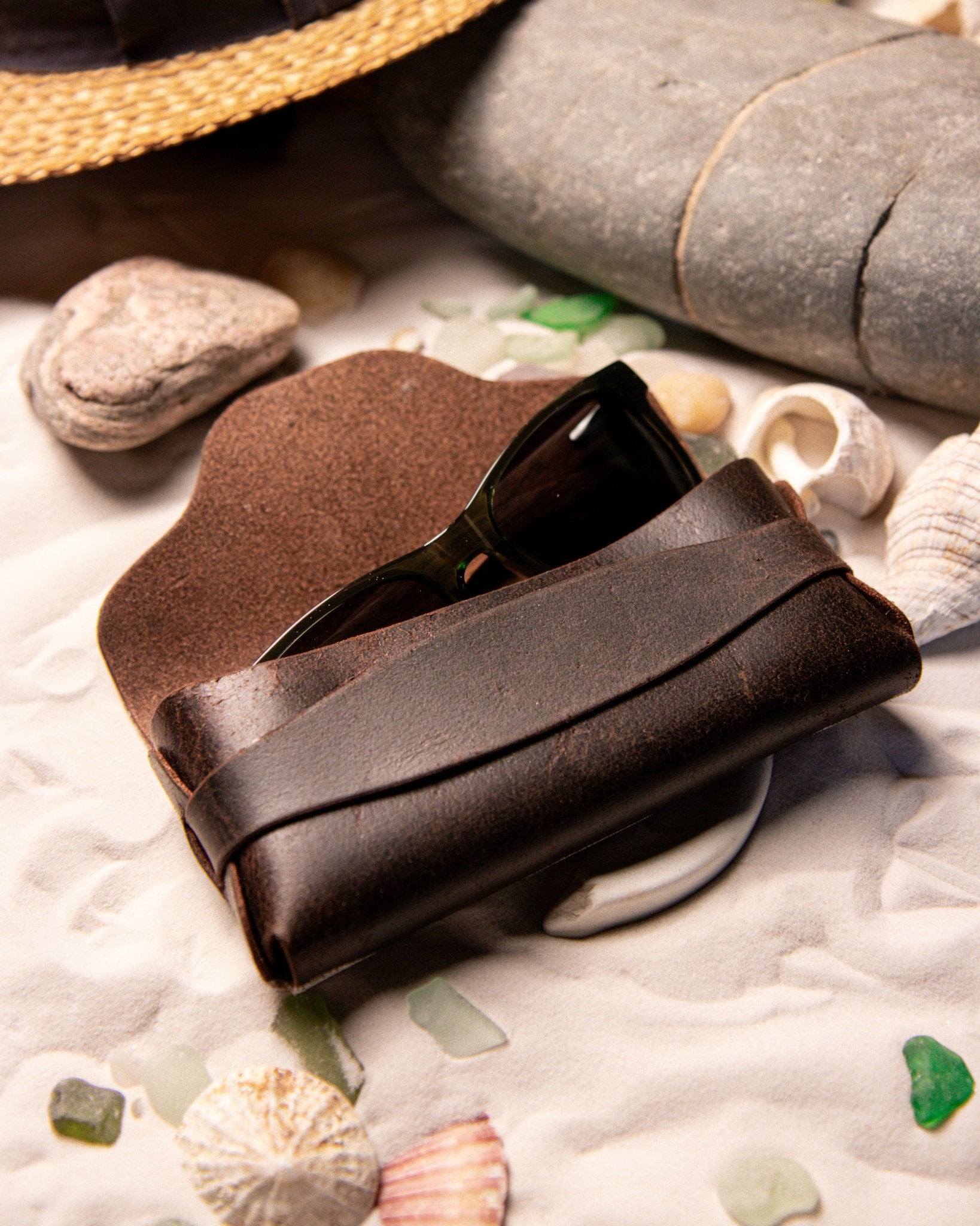 Brown leather sunglasses case with sunglasses on a beach setting with stones and shells.