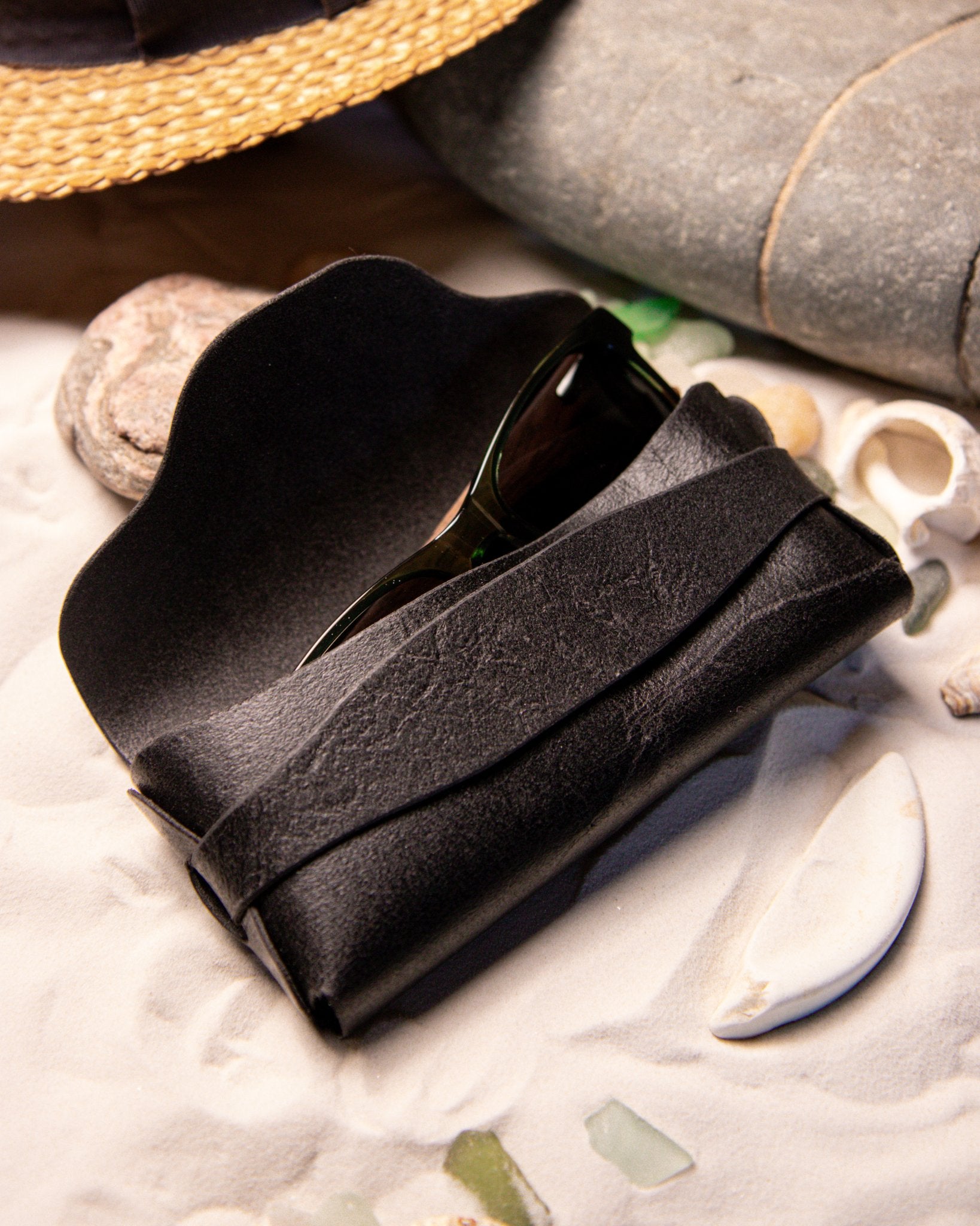 Black sunglasses in a eyewear case on a textured surface with stones and shells.